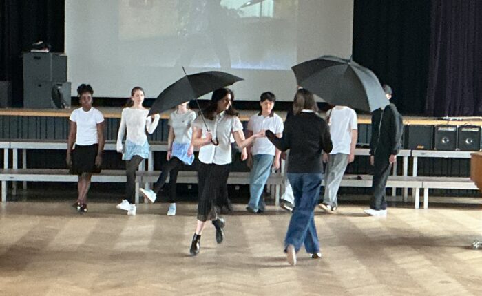 Eine junge Frau mit einem Regenschirm tanzt auf einer Bühne, umgeben von anderen Personen.