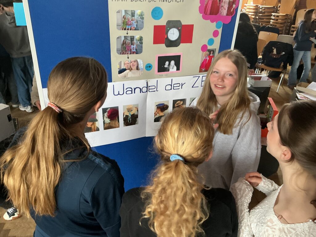 Eine lächelnde Schülerin mit langen, blonden Haaren steht vor einem Plakat.