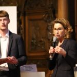 Eine Frau mit lockigem Haar und Brille spricht ins Mikrofon, während ein junger Mann zusieht.