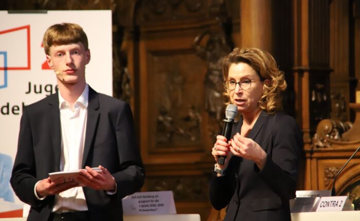 Eine Frau mit lockigem Haar und Brille spricht ins Mikrofon, während ein junger Mann zusieht.
