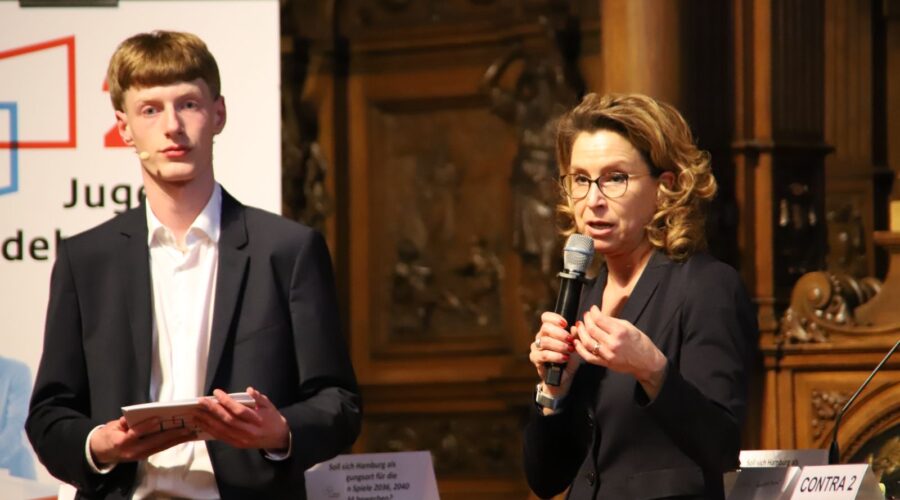 Eine Frau mit lockigem Haar und Brille spricht ins Mikrofon, während ein junger Mann zusieht.