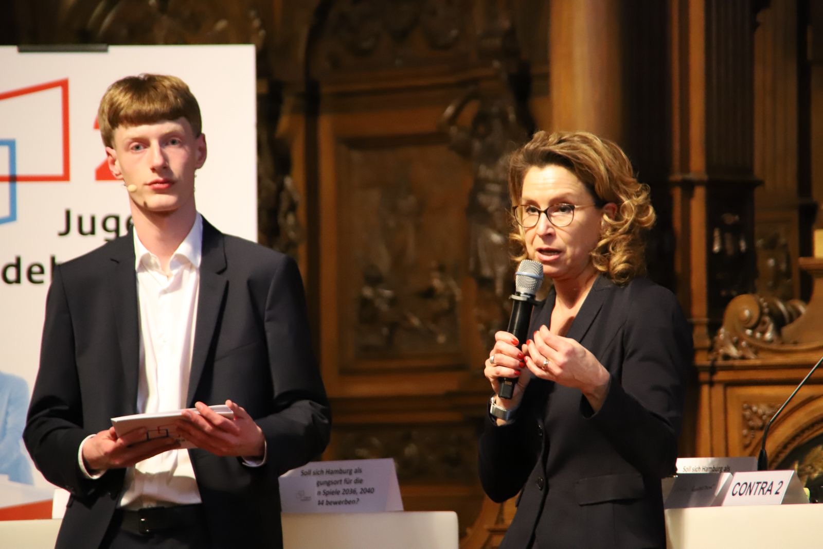 Eine Frau mit lockigem Haar und Brille spricht ins Mikrofon, während ein junger Mann zusieht.