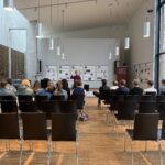 Ein Mann steht vor einer Gruppe von Personen in einem hellen, modernen Raum.