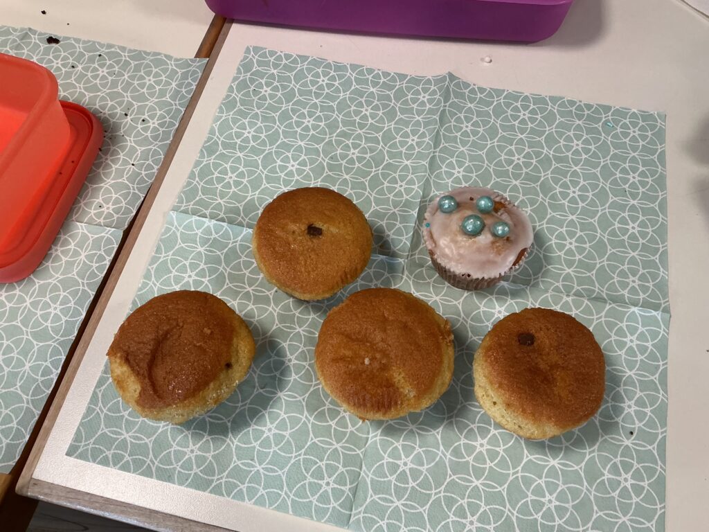 Fünf Muffins auf einem dekorativen Papier, einer mit Zuckerglasur und blauen Perlen.