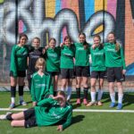 Die Gruppe von neun jungen Fußballspielerinnen posiert vor einer bunten Wand mit Graffiti.