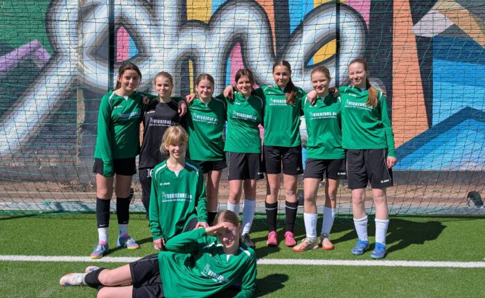 Die Gruppe von neun jungen Fußballspielerinnen posiert vor einer bunten Wand mit Graffiti.