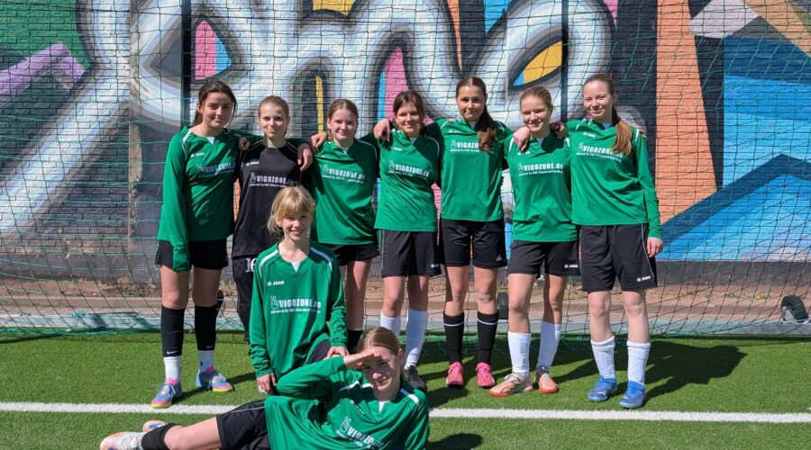 Die Gruppe von neun jungen Fußballspielerinnen posiert vor einer bunten Wand mit Graffiti.