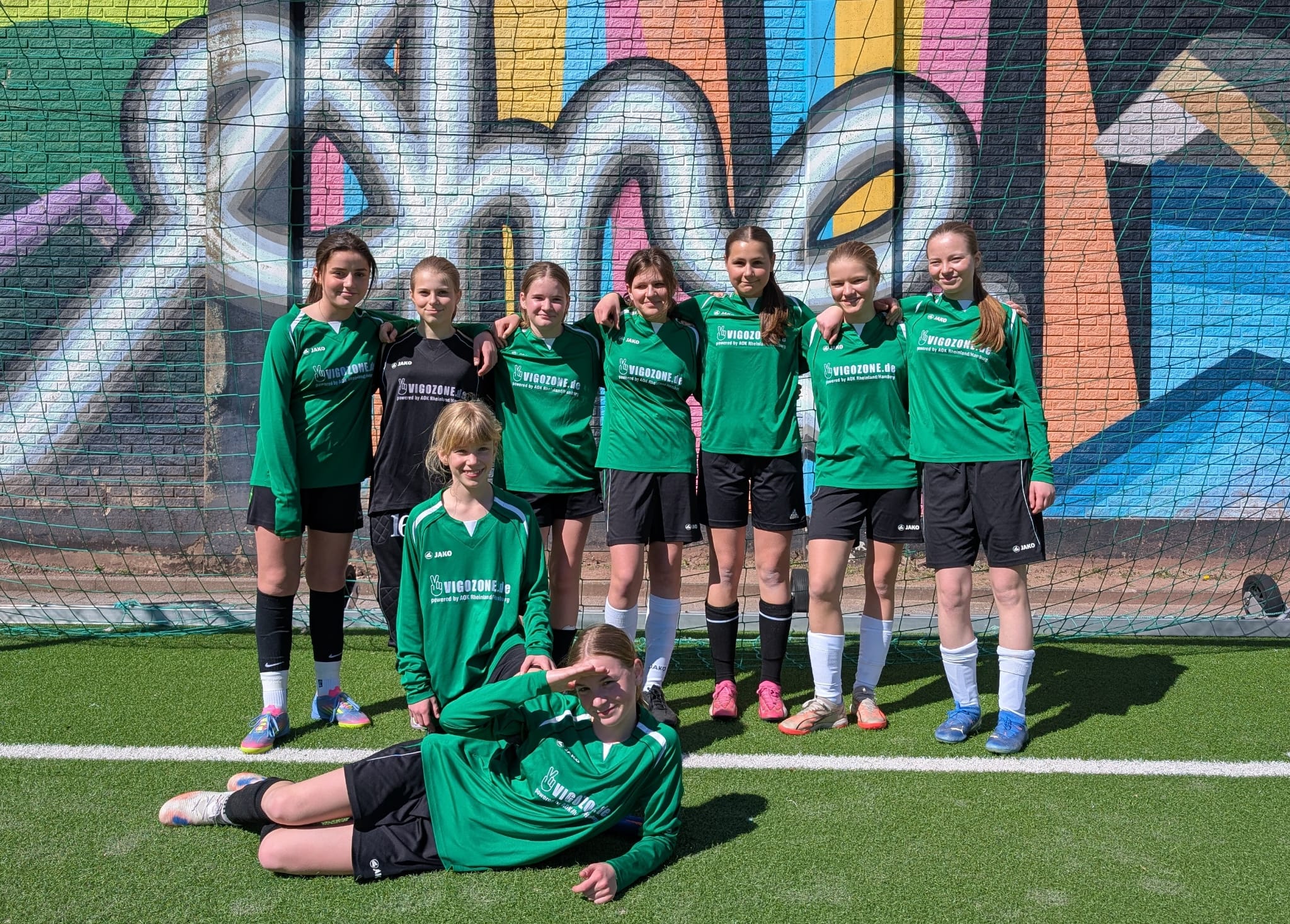 Die Gruppe von neun jungen Fußballspielerinnen posiert vor einer bunten Wand mit Graffiti.