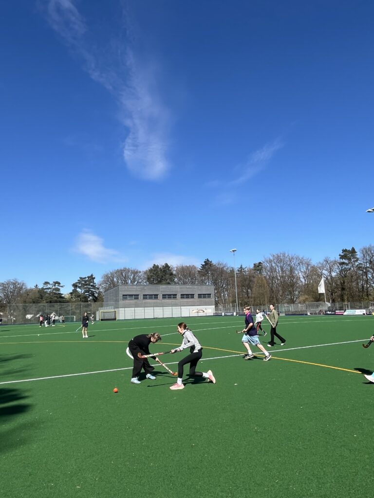 Zwei Spielerinnen im Wettkampf um den Ball auf einem Hockeyfeld unter blauem Himmel.