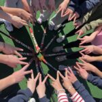 Mehrere Hände formen ein V-Zeichen, umgeben von Hockeyschlägern auf grünem Untergrund.