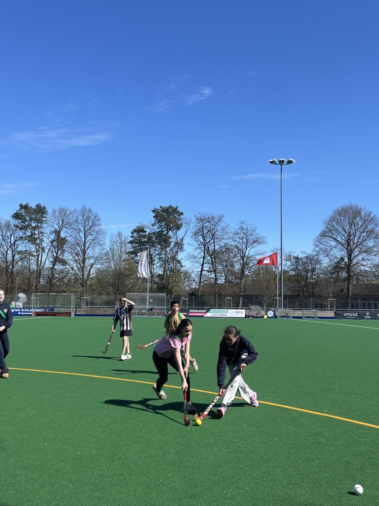 Zwei Personen spielen Hockey auf einem kunstrasengrünen Spielfeld bei klarem Himmel.