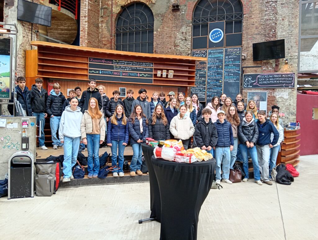 Eine Gruppe von Jugendlichen posiert lächelnd vor einer vorderen Bühne in einem Veranstaltungsort.