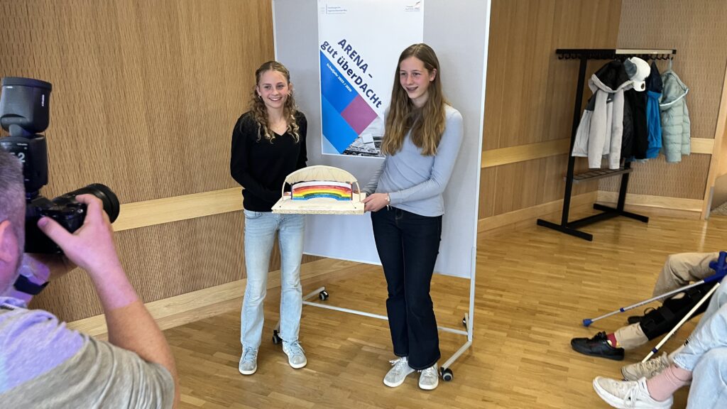 Zwei junge Frauen halten ein Modell mit einem Regenbogen und lächeln in die Kamera.