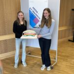 Zwei junge Frauen halten ein Modell mit einem Regenbogen und lächeln in die Kamera.