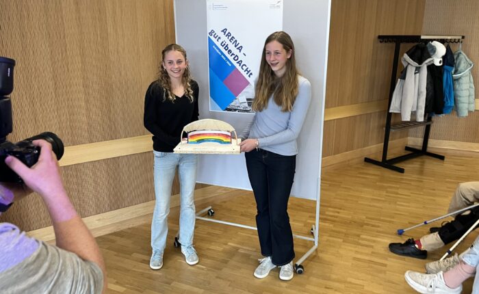 Zwei junge Frauen halten ein Modell mit einem Regenbogen und lächeln in die Kamera.