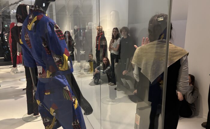 Dunkelhäutige Person mit blauem, gemustertem Outfit steht vor einer Glasscheibe.