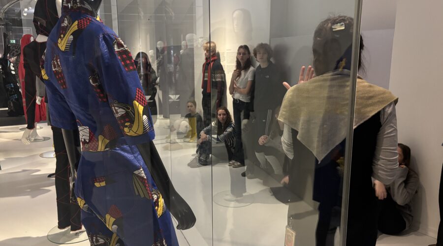 Dunkelhäutige Person mit blauem, gemustertem Outfit steht vor einer Glasscheibe.