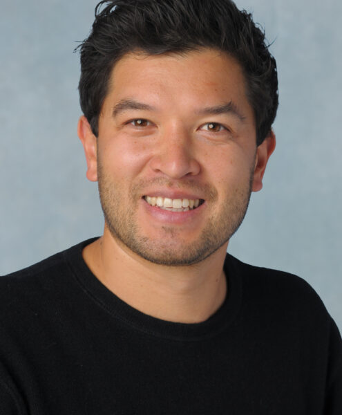 Porträt eines Mannes mit dunklem Haar, lächelnder Gesichtsausdruck, schwarzem Pullover, hellem Hintergrund.