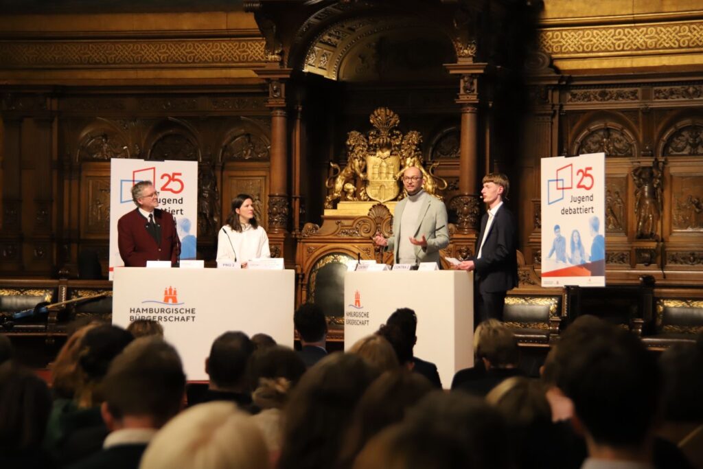 Vier Personen stehen an Pulten und diskutieren in einem historischen Saal.