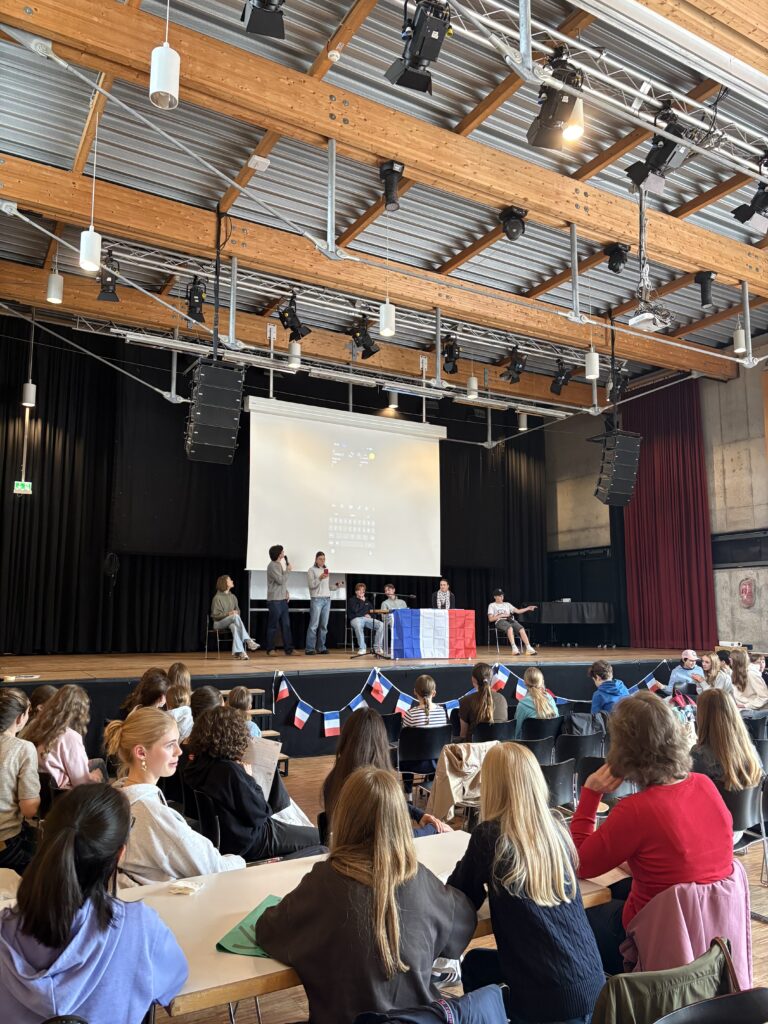 Schülerinnen und Schüler sitzen im Auditorium, während eine Diskussion auf der Bühne stattfindet.