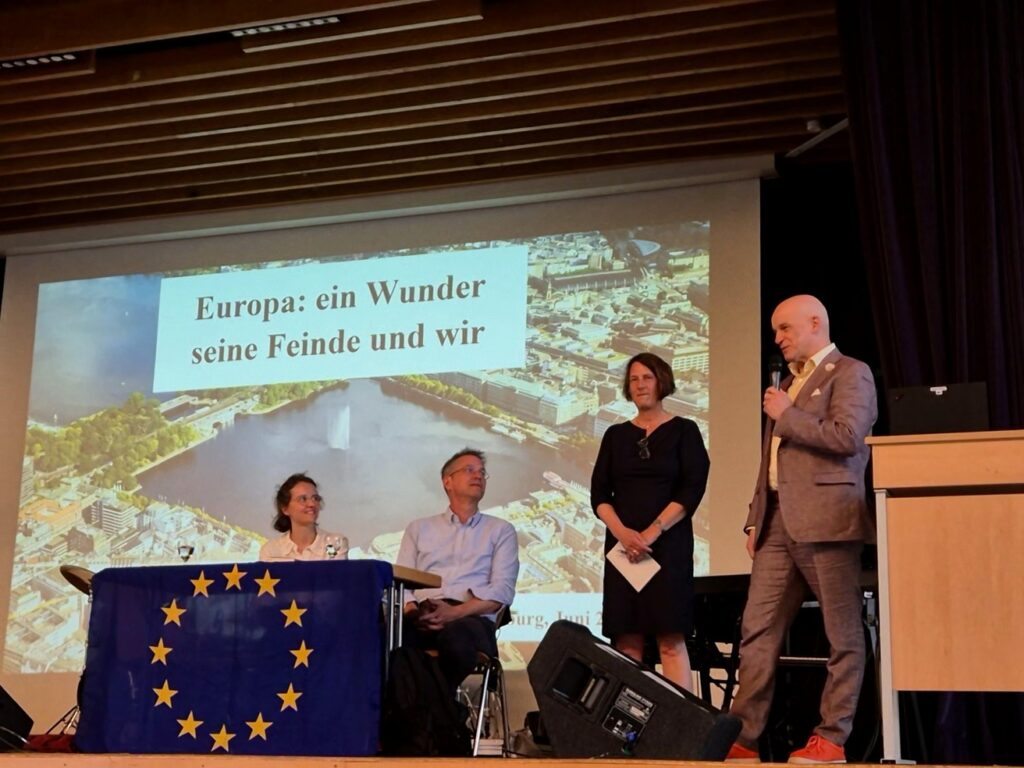 EinModerator spricht am Mikrofon vor einer Präsentation mit dem Titel „Europa: ein Wunder seine Feinde und wir“.
