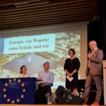 EinModerator spricht am Mikrofon vor einer Präsentation mit dem Titel „Europa: ein Wunder seine Feinde und wir“.