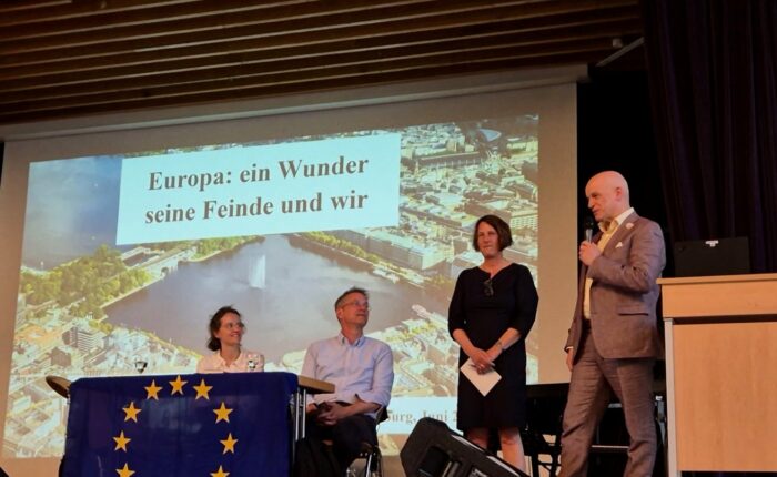 EinModerator spricht am Mikrofon vor einer Präsentation mit dem Titel „Europa: ein Wunder seine Feinde und wir“.
